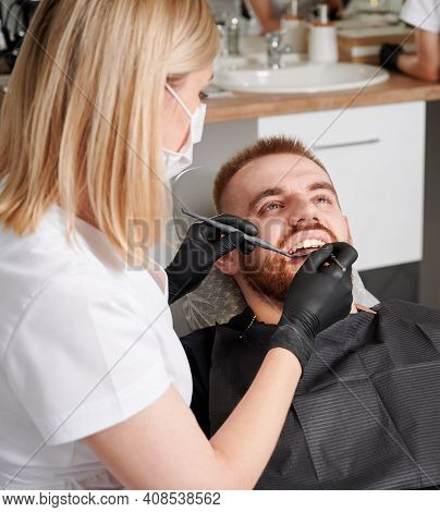 Dentist Examining Patient Teeth With Dental Explorer And Mirror. Bearded Young Man Lying In Dental C