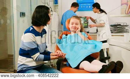 Pediatric Dentist Checking Teeth X-ray Of Kid Patient Talking With Man Assistant In Background. Sad 