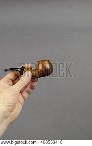 Hand Holds A Smoking Pipe On A Gray Background. Bend A Briar Or Heather Root Smoking Pipe. Tobacco S