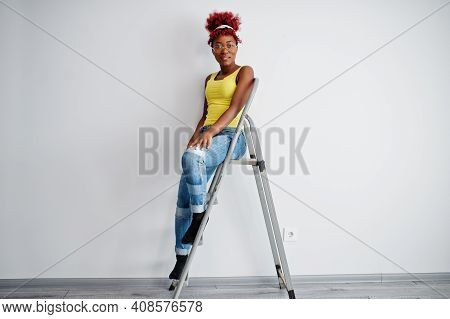 African American Woman In Yellow Singlet And Eyglasses Against White Wall Stand Om Iron Ladder.