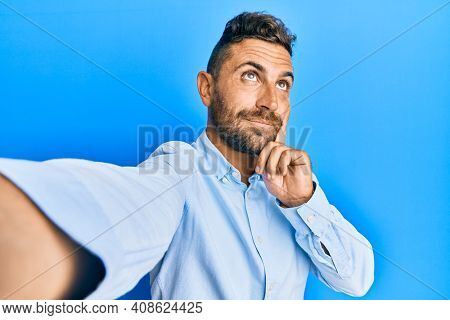 Handsome man with beard taking a selfie photo with smartphone serious face thinking about question with hand on chin, thoughtful about confusing idea 