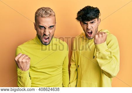 Homosexual gay couple standing together wearing yellow clothes angry and mad raising fist frustrated and furious while shouting with anger. rage and aggressive concept. 