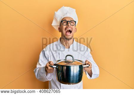 Bald man with beard wearing professional cook holding cooking pot angry and mad screaming frustrated and furious, shouting with anger looking up. 