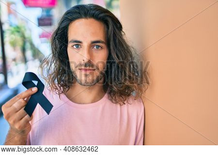 Young hispanic man with serious expression holding mouring black ribbon leaning on the wall.