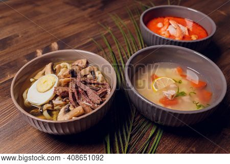 Set Of Serving Asian Soups On Gray Background Top View. Tom Yum Soup, Eel Fish Soup And Ramen Noodle