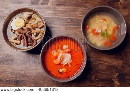 Set Of Serving Asian Soups On Gray Background Top View. Tom Yum Soup, Eel Fish Soup And Ramen Noodle