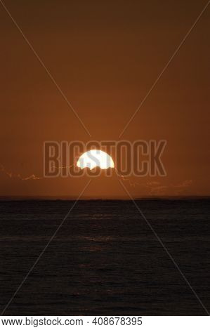 A Calm Ocean Forefront With The Orange Sun Descending Over The Clouds In The Background.