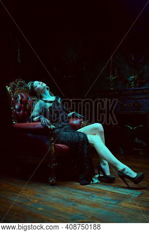 Beautiful woman in an elegant evening dress in an armchair in a rich classic interior. Clothing, makeup and hairstyle in the style of the 1920s.