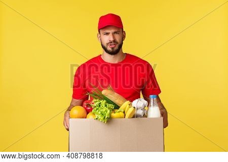 Online Delivery, Shopping And Food Order Concept. Doubtful And Upset Courier In Red Uniform Cap And 