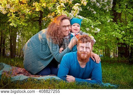 Young Family With Kid Having Picnic Outdoors Near Apple Tree With White Flowers. Parents With One Ch