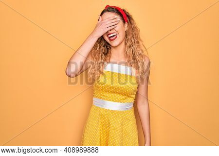 Beautiful blonde pin-up woman with blue eyes wearing diadem standing over yellow background smiling and laughing with hand on face covering eyes for surprise. Blind concept.