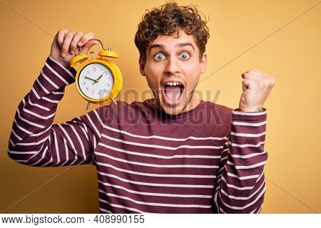 Young blond man with curly hair holding alarm clock standing over isolated yellow background screaming proud and celebrating victory and success very excited, cheering emotion