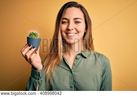 Young beautiful blonde woman with blue eyes holding small cactus pot over yellow bckground with a happy face standing and smiling with a confident smile showing teeth