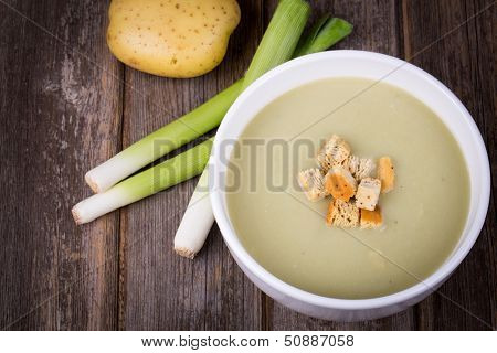 A bowl of leek and potato soup with bread croutons, over old wood table with fresh leeks and potatoes alongside