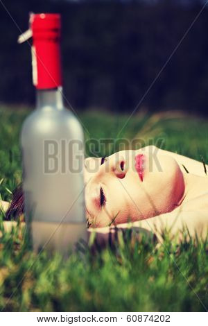 Drunk young woman is sleeping on the grass next to bottle of alcohol in sunny day.