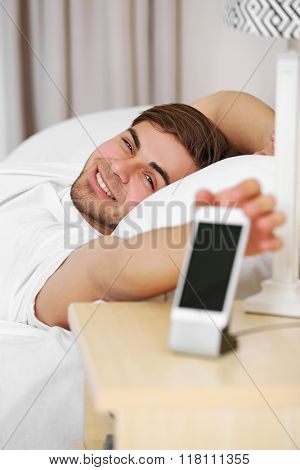 Beautiful young man waking up with mobile alarm clock