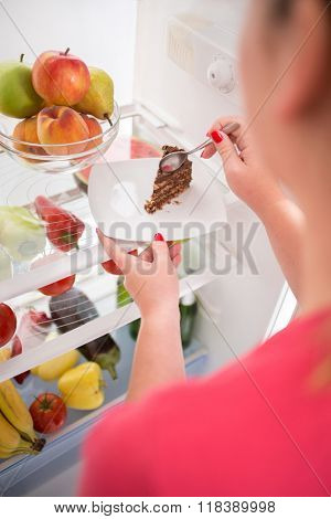 Close up young woman on diet stealing from fridge piece of nice chocolate cake