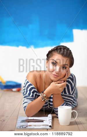 Portrait of female painter lying on floor near wall after paintingand holding a cup