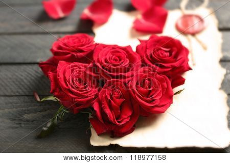Fresh red roses buds in shape of heart with blank present card on wooden background