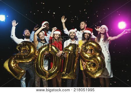New 2018 year is coming. Group of cheerful young people in Santa hats holding gold colored number balloons, showered with confetti. Christmas postcard mokup, celebration concept