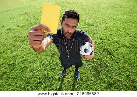 High angle view of confident mixed race soccer referee showing yellow card to camera while standing at spacious football pitch illuminated with sunbeams