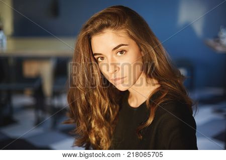 Horizontal picture of beautiful young European dark haired female student wearing black dress looking at camera with serious expression while sitting at university canteen during lunch break