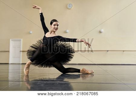 Attractive ballerina posing in the dance hall. Her arms outstretches to the sides, right leg is on the toe. She wears a black dance wear with dark tutu and light pointe shoes. Horizontal.