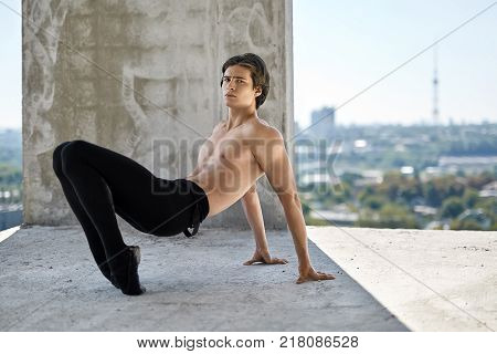 Topless ballet dancer is posing on the concrete floor of the unfinished building on the cityscape background. He wears a black dance pants and pointe shoes. Guy leans on his hands and toes.