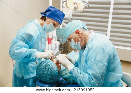 Male Old Professional Dentist Surgeon In Uniform And Female Assistant Helps To Performing Operation