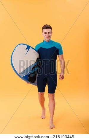 Full lenth photo of cheerful guy in swimsuit holding surfboard while walking and looking at camera, isolated on yellow background