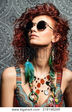 Close-up portrait of a beautiful bright woman with curly foxy hair in sunglasses, earrings and beads. Ethnic style in accessories.