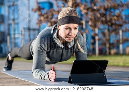 High-angle view of a fit young woman, watching a motivational video on tablet PC while exercising the forearm plank position on a mat outdoors in the park