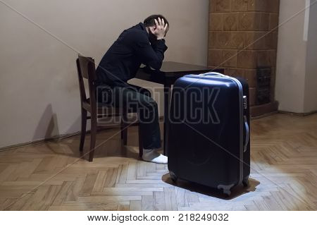 A young man sits bent over the table his head in his hands near the table is a large travel bag