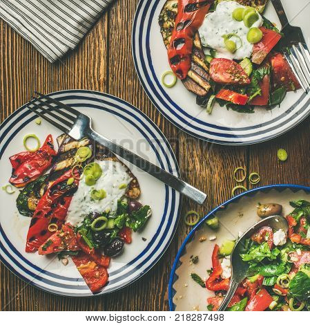 Flat-lay of healthy dinner table setting. Green salad, grilled vegetables with yogurt sauce, pickled olives on plates, lemon water over wooden background, top view, square crop. Vegan food concept