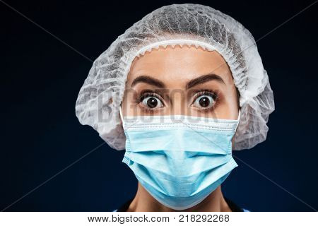 Portrait of shocked doctor in medical cap and mask looking camera isolated over black