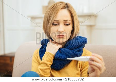 Having a cold. Attractive sad young blond woman feeling down in the mouth and holding a thermometer and having a blue warm scarf on while sitting on the sofa