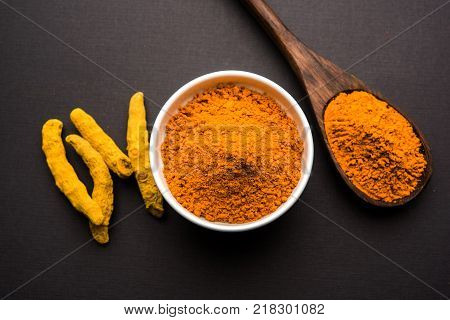Turmeric powder in ceramic bowl with raw dried turmeric over plain background