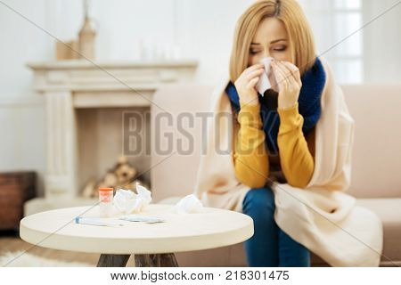 Feeling ill. Ill young blond woman feeling bad and blowing her nose while having a blanket on her shoulders and sitting on the couch and table with pills in front of her