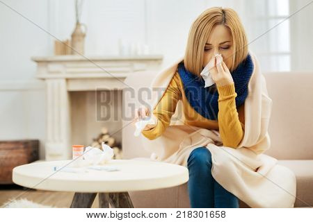 Runny nose. Ill young blond woman having fever and blowing her nose while having a blanket on her shoulders and sitting on the couch with her eyes closed and table with pills in front of her