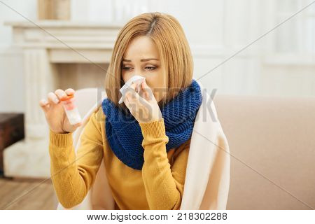 Having disease. Pretty ill young blond woman having fever and blowing her nose and holding medication while having a blanket on her shoulders and sitting on the couch