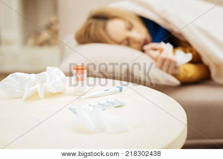 Taking a nap. Sick young blond woman feeling under the weather and sleeping on bed and a table with napkins and medications standing in front of the couch