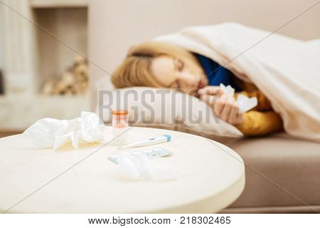 Sleeping. Ill young blond woman feeling under the weather and sleeping on bed and a table with napkins and medications standing in front of the couch