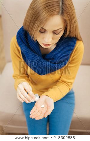 Recovery. Unhappy young blond long-haired woman wearing a scarf jeans and sweater and sitting on the couch and holding pills