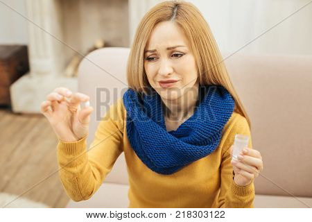 Yuck. Sick young blond long-haired woman wearing a scarf jeans and sweater and sitting on the couch while holding a pill and looking at it with disgust