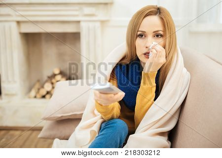 Sick leave. Beautiful cheerless young dark-eyed woman having runny nose and feeling unwell and sitting on the couch and holding remote control