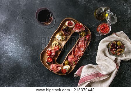 Tapas or bruschetta variety. Bread with ham prosciutto, sun dried tomatoes, olive oil, olives, pepper on decorative wood plate with glass of red wine over dark texture background. Top view with space