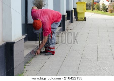 Cleaning of a city street - the worker the yard keeper in a uniform collects garbage by hands near a building behind it there is a trash can.