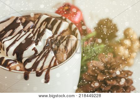 Hot chocolate in white cup topping with marshmallow and dark chocolate sauce. Delicious hot chocolate or cacoa on wood table in close up view copy space with Christmas theme and snowfall background. Concept to present hot chocolate for Christmas drink.