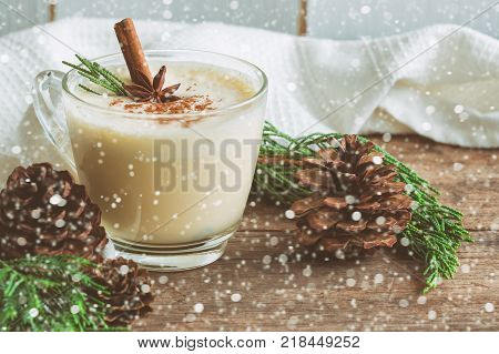 Eggnog traditional drink for Christmas celebration party. Homemade eggnog in cup topped with ground nutmeg and cinnamon stick top view with copy space in Christmas theme decoration and snowfall effect background. Delicious eggnog ready to served.