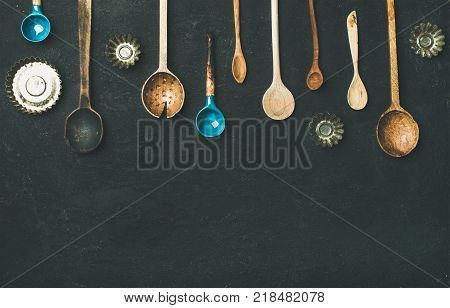 Flat-lay of various old vintage kitchen spoons and baking tin molds over black stone background, top view, copy space. Rustic cooking concept
